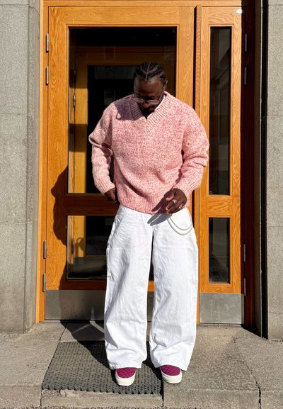 Hombre con el pelo trenzado que lleva un suéter de punto rosa, pantalones anchos blancos y zapatillas con estampado, está delante de una puerta de madera sobre escalones de piedra.