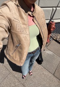 Casaco bomber de camurça em tom areia com "EASY" bordado no peito, combinado com uma camisola de alças de malha em verde claro, jeans de ganga e sandálias pretas.