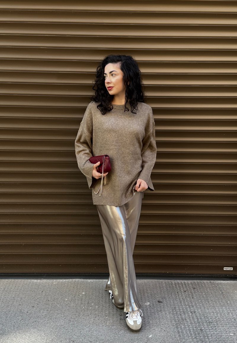 Pull beige surdimensionné à manches évasées, associé à un pantalon large argenté brillant. Tenant une pochette bordeaux avec une bandoulière en chaîne.