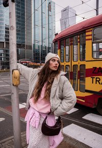 Een grijze pufferjack over een roze hoodie, een witte kanten rok en een roze shirt geknoopt in de taille. Een bruine tas en een grijs gebreid mutsje. Tram op de achtergrond.