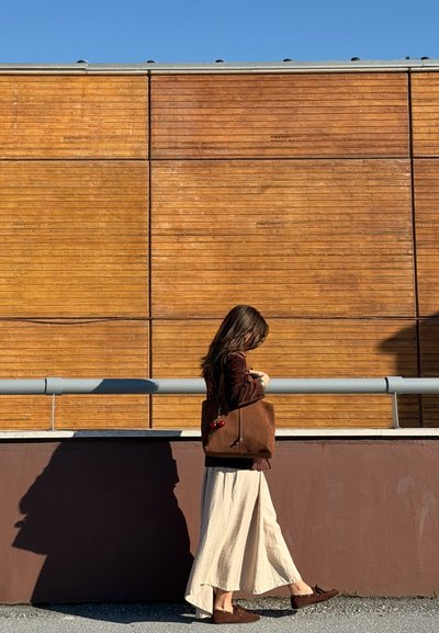 Mujer con chaqueta marrón y falda beige camina junto a una pared de madera, llevando un gran bolso marrón con un llavero de cereza bajo un cielo azul claro.