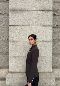 Vrouw met donker haar in een zwart jasje met knopen, zijwaarts staand tegen een lichtgrijze stenen muur, met een donker gevlochten tas in haar hand.