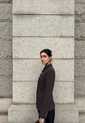 Mujer de cabello oscuro con blazer negro abotonado, de pie de lado contra una pared de piedra gris clara, sosteniendo un bolso tejido oscuro.