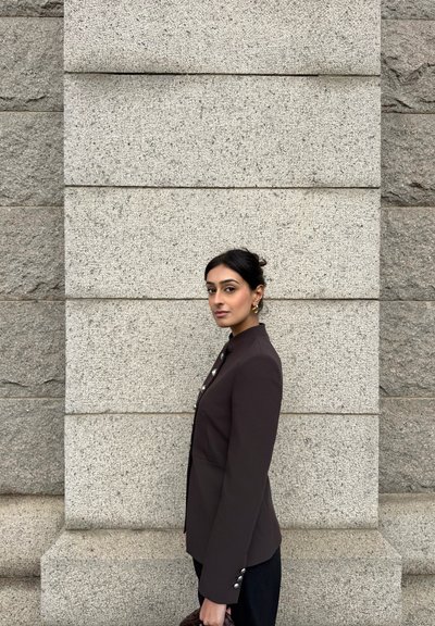 Mujer de cabello oscuro con blazer negro abotonado, de pie de lado contra una pared de piedra gris clara, sosteniendo un bolso tejido oscuro.