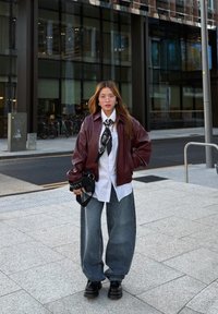 Burgundy bőr bomber dzseki fehér ing felett, fekete mintás nyakkendő és széles kék farmer; fekete platform cipők zárják le a megjelenést.