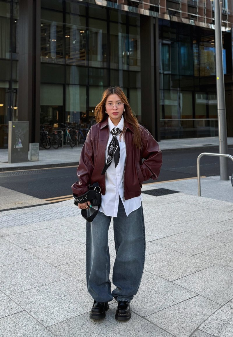 Burgundia nahast bomber-jakk valge särgi, musta mustriga lipsu ja laia sinise teksapüksiga; välimuse lõpetavad mustad platvormisaapad.