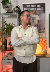 Strikket kremfarget vest over en stripete skjorte, med en avslappet passform. Bakgrunnen inkluderer planter, dekor og en mykt opplyst globelampe.