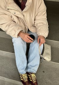 Μπεζ ζακέτα καμπαρντίνα oversized, ανοιχτό μπλε τζιν, μπορντό και κίτρινες αθλητικές κάλτσες, κρατώντας ένα μαύρο φλιτζάνι, με μια μικρή μπεζ τσάντα στο πλάι.