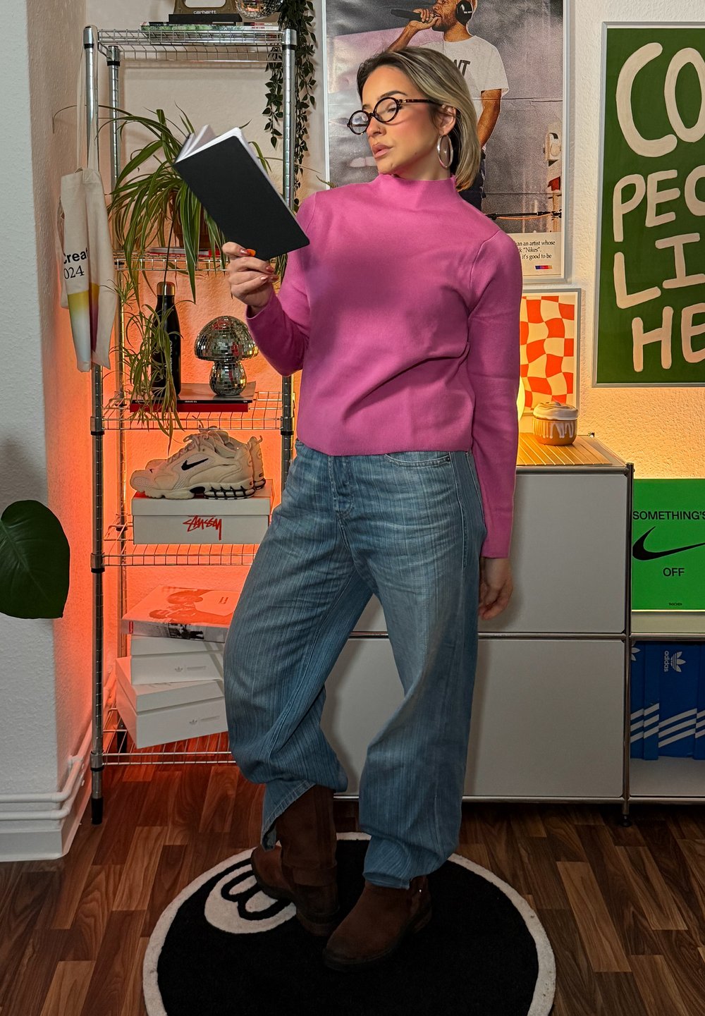 Pulover roz cu guler înalt, blugi largi de culoare albastru deschis, cizme maro, ținând un caiet negru, într-o cameră colorată cu plante și decorațiuni.