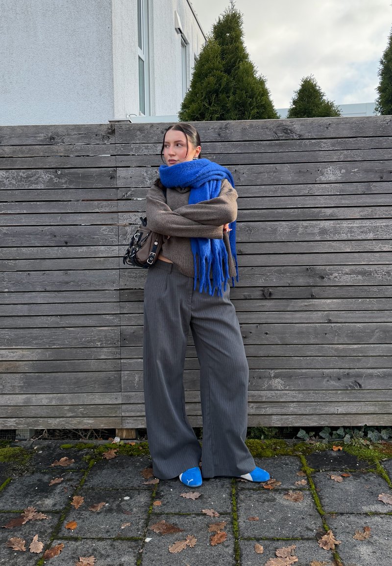 Mulher de pé com os braços cruzados, vestindo calças cinzentas, camisola castanha, cachecol azul brilhante e sapatos, em frente a uma cerca de madeira com árvores ao fundo.