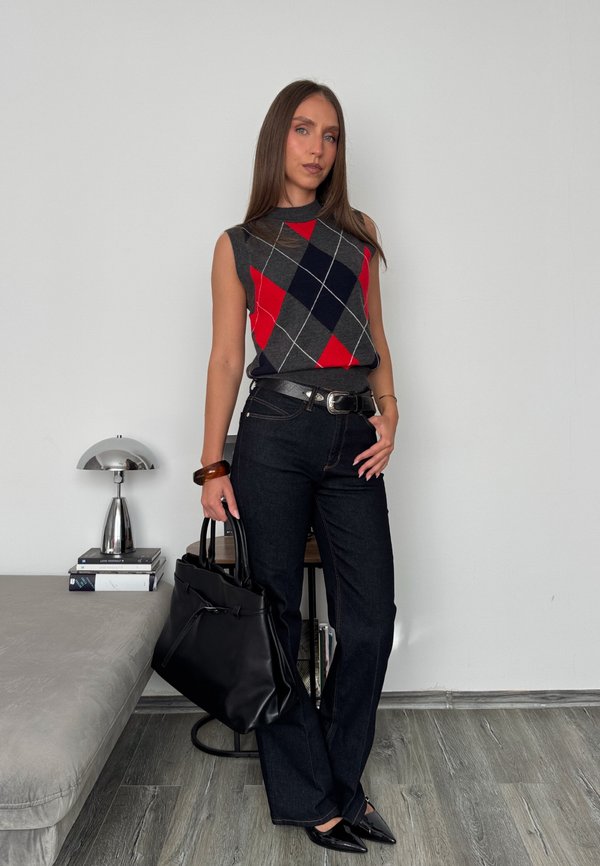 Jeune femme portant un pull sans manches à motif losanges et un jean noir, tenant un sac à main noir, debout dans une pièce moderne avec une lampe et des livres.