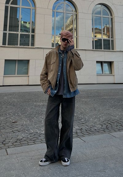 Joven con gafas de sol, chaqueta marrón claro, vaqueros superpuestos y pantalones anchos oscuros, de pie sobre pavimento adoquinado cerca de un edificio moderno con ventanas arqueadas.