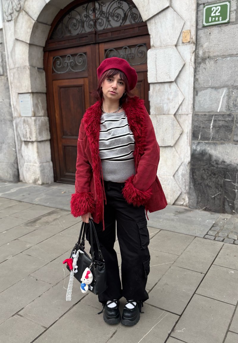 Long manteau en velours côtelé rouge avec bordure en fausse fourrure, pull rayé gris, et pantalon cargo noir. Sac à main noir avec des breloques, chaussures noires épaisses.