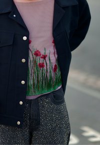Svart jacka med silverknappar över en ljust lila topp med röd blommig broderi, ihopparad med svarta byxor prydda med silverstift.