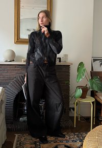 Črni pleteni pulover z visokim ovratnikom, kombiniran z širokimi črnimi hlačami. V ozadju je teksturiran kamnit kamin in sobne rastline.