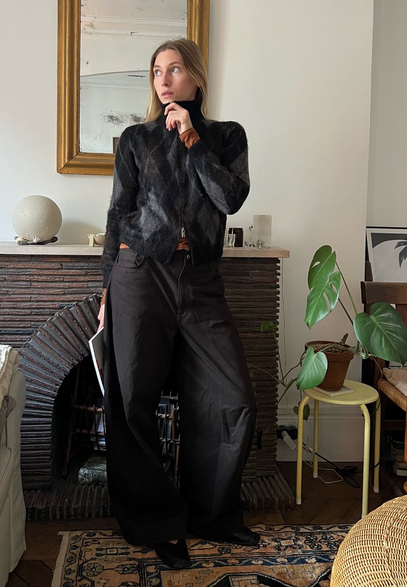Camisola preta de malha com gola alta, combinada com calças largas pretas. O fundo apresenta uma lareira de pedra texturizada e plantas interiores.