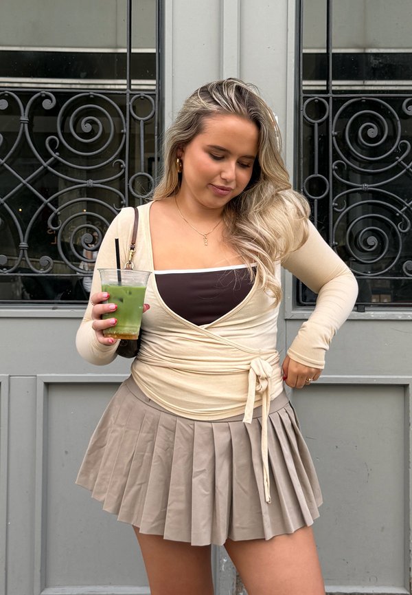 Jonge vrouw met lang blond haar draagt een beige wikkeltop en geplooide rok, houdt een groen koud drankje met rietje vast voor een decoratieve deur.