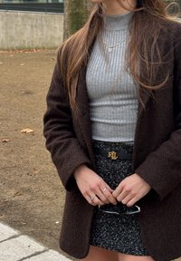 Γυναίκα που φοράει καφέ παλτό με υφή, γκρι ζιβάγκο με ριμπ ύφανση, μαύρη τουίντ φούστα με ζώνη RL, κρατώντας γυαλιά ηλίου με πολλά δαχτυλίδια στα δάχτυλα, σε εξωτερικό χώρο.