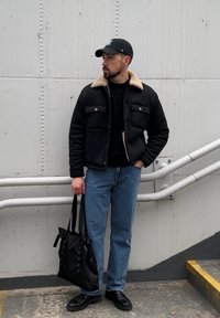 Veste noire avec col en fourrure marron clair, pull noir, jean bleu et baskets noires. Tenant un sac fourre-tout noir, debout contre un mur gris.