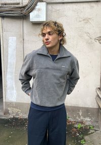 Šedý fleecový pulovr s vysokým límcem, předním švem a malým logem na hrudi. Nošený s tmavými volnými kalhotami.