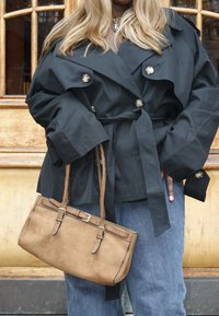 Čierny, nadrozmerný trenčkot s veľkými gombíkmi a opaskom, doplnený hnedou semišovou kabelkou so strapcom a detailmi na pracke.