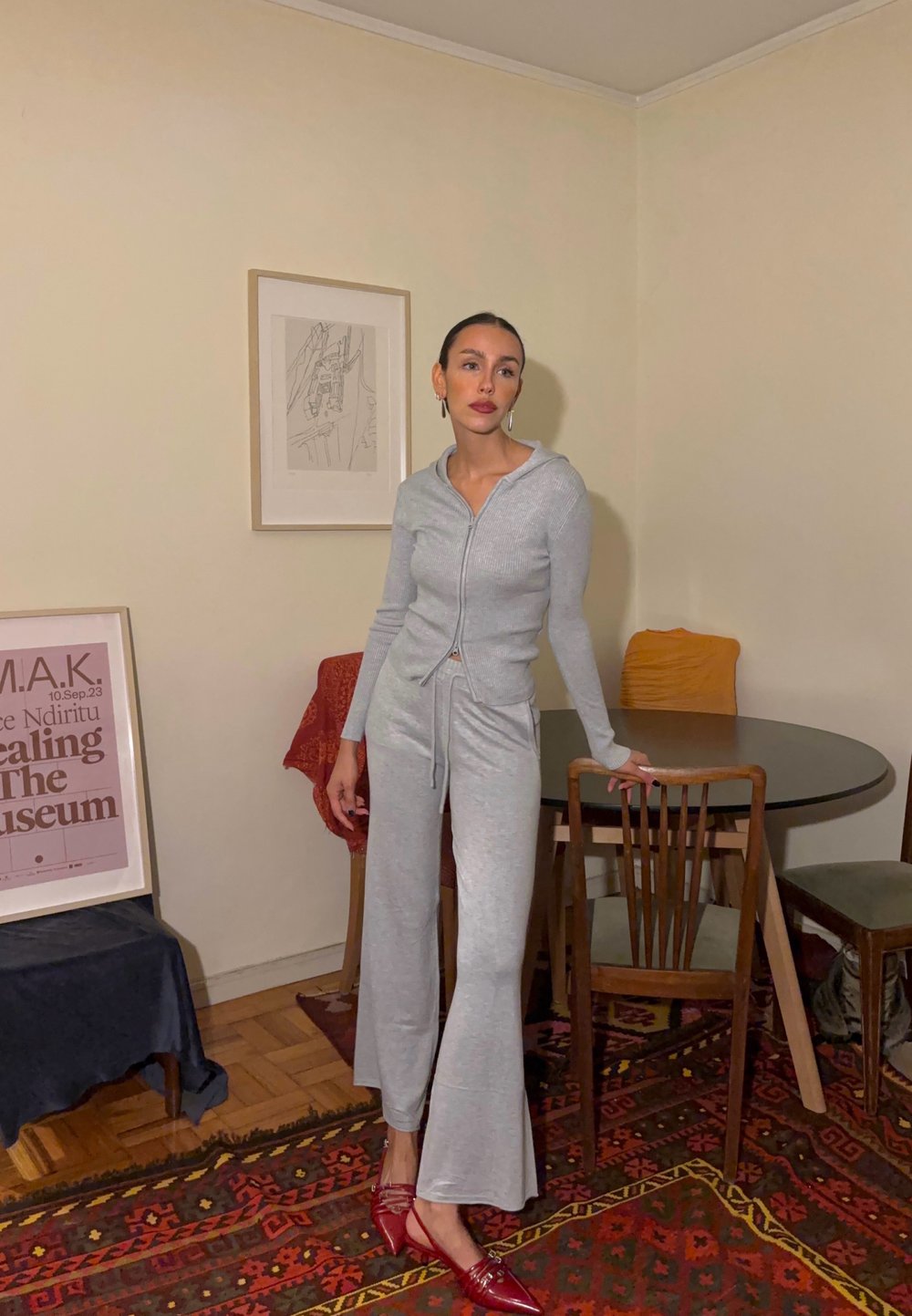 Pelēkas ribotas brīvdienu apģērbu komplekts ar rāvējslēdzēja kapuci, plašajām biksēm un sarkanām smailēm, izvietots mājīgā iekštelpā ar koka mēbelēm.