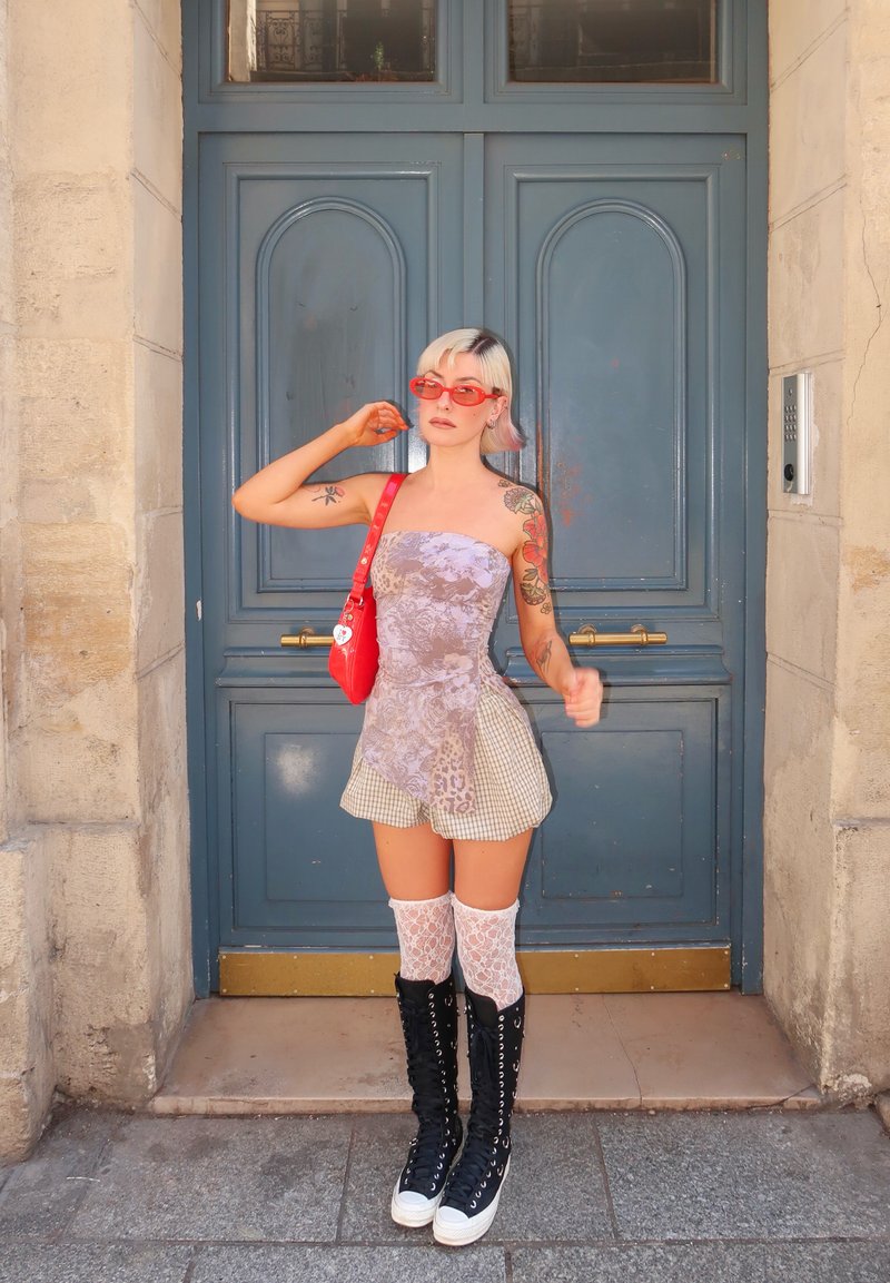 Woman with short blonde hair wearing a tube top dress, red sunglasses, red bag, lace knee-high socks, and black high-top boots stands by a blue door.
