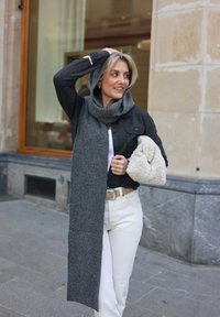 Lange grijze wollen sjaal die over een zwarte jas valt, gecombineerd met een witte broek en een pluizig accessoire in crèmekleur.