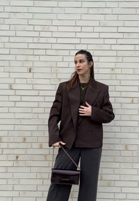 Femme aux cheveux foncés portant un blazer marron et un pantalon gris, tenant un sac à main foncé, debout contre un mur en briques blanches, regardant sur le côté.