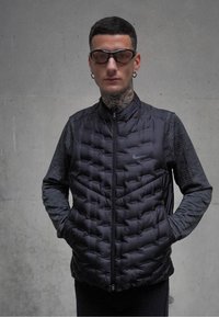 Veste sans manches noire matelassée avec une texture matelassée, fermeture éclair à l'avant et col montant. Sous-vêtement noir à manches longues avec un motif texturé.
