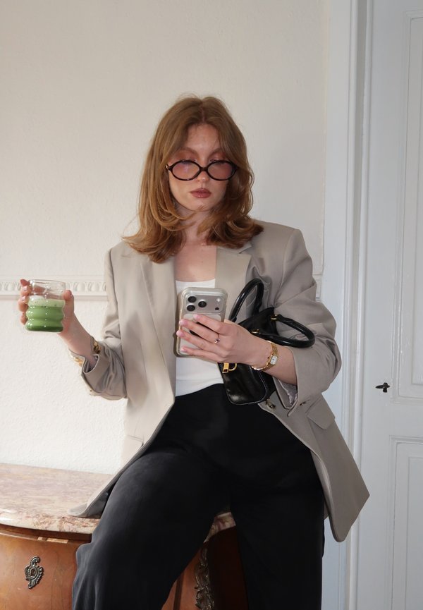 Femme aux cheveux rouges et lunettes tenant une boisson verte et un téléphone, portant un blazer beige, un haut blanc et un pantalon noir, assise sur des meubles en bois.