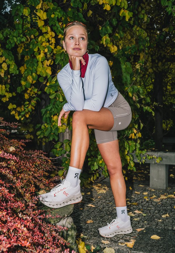 Sportkleding omvat een lichtgrijs longsleeve shirt met een hoge hals, beige shorts en witte sneakers met rode accenten.