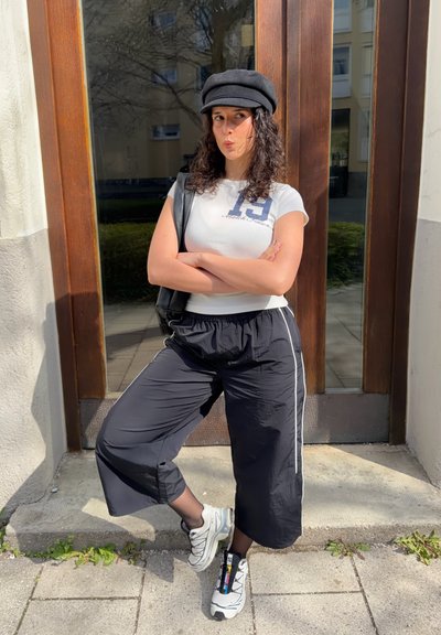 Joven mujer con gorra negra, camiseta blanca ajustada y pantalones negros sueltos, de pie con los brazos cruzados, una pierna doblada, frente a una puerta de madera.