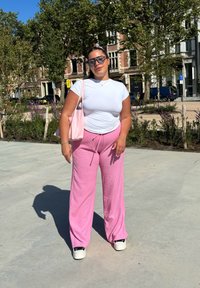 Haut blanc ajusté à manches courtes, pantalon large texturé rose, et baskets noires. Tenant un sac fourre-tout rose clair.