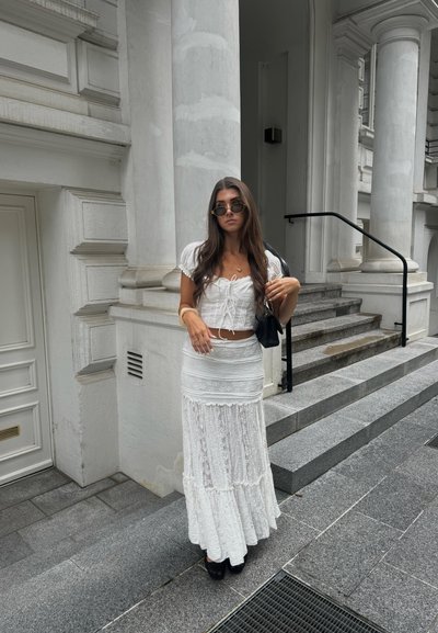 Conjunto blanco de dos piezas que presenta un top ajustado con un lazo en la parte delantera y una falda larga y acampanada. Los accesorios incluyen gafas de sol redondas y un bolso negro pequeño.