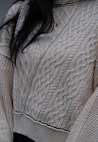 Maglione a maglia cables di colore avorio con motivi testurizzati, colletto alto e accento nero lungo l'orlo a costine. Materiale morbido e caldo.