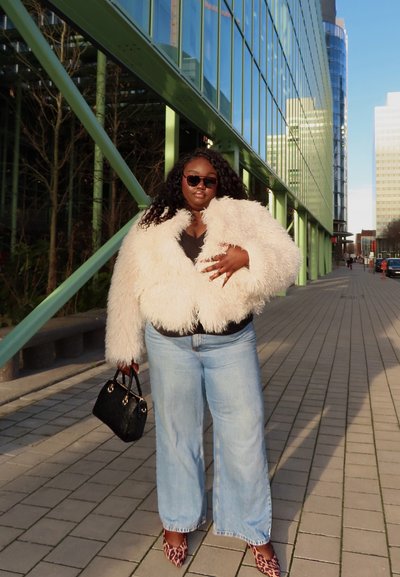 Mujer con chaqueta blanca esponjosa, vaqueros azul claro, zapatos de estampado de leopardo, gafas de sol y un bolso negro, en una calle de la ciudad.
