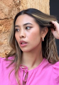 Μοντέλο με μακριά, κυματιστά μαλλιά, φορώντας ροζ πουκάμισο με φουσκωτά μανίκια και λεπτομέρεια με κομψό δέσιμο, παρέα με ασημένια σκουλαρίκια κρίκους και φυσικό μακιγιάζ.