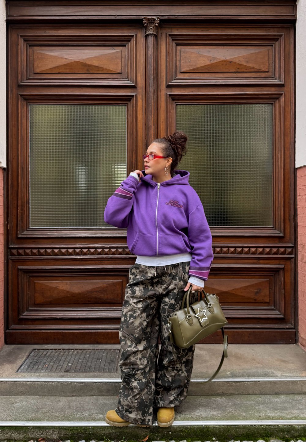 Sweat à capuche violet à fermeture éclair avec poche avant, pantalon ample à motif camouflage, sac à main olive avec détail étoile, et chaussures beige. Fond de porte en bois texturé.