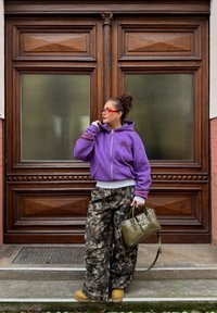 Sweat à capuche violet à fermeture éclair avec poche avant, pantalon ample à motif camouflage, sac à main olive avec détail étoile, et chaussures beige. Fond de porte en bois texturé.