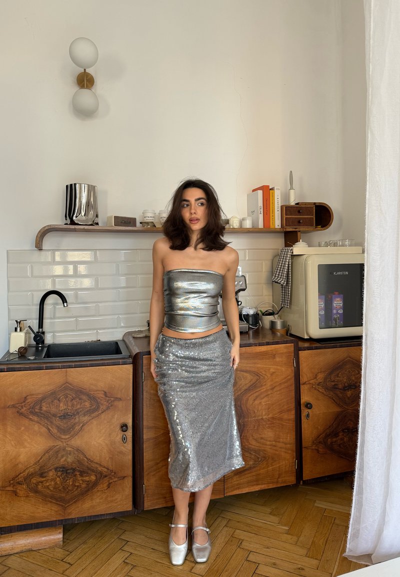 Top argentato senza spalline e gonna argentata con paillettes; texture e scintillante, abbinati a scarpe argentate lucide, su uno sfondo di armadietti in legno.