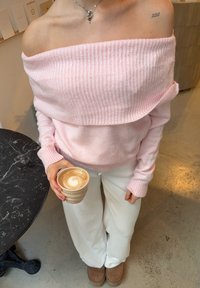 Maglione rosa a coste indossato leggermente sceso sulle spalle, abbinato a pantaloni bianchi larghi in velluto a coste; tiene in mano un latte in una tazza beige dalla superficie strutturata.