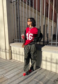 Rød idrettsdrakt med nummer 46, svart oversized jakke, camo cargo bukser og røde slip-on sko. Holder en klar drink i en kopp.