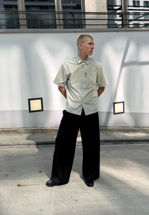 Personne aux cheveux blonds courts portant une chemise légère à manches courtes boutonnée et un pantalon large noir, debout contre un mur blanc à l'extérieur.