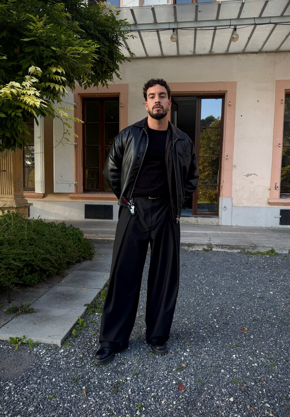 Uomo con capelli ricci e barba sta all'aperto indossando una giacca di pelle nera, pantaloni larghi neri e scarpe nere vicino a un edificio e della vegetazione.