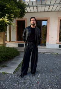 Uomo con capelli ricci e barba sta all'aperto indossando una giacca di pelle nera, pantaloni larghi neri e scarpe nere vicino a un edificio e della vegetazione.