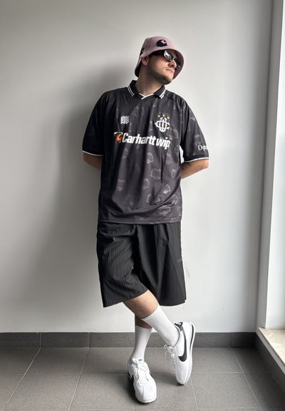 Jersey negro de manga corta con ribetes y logo blancos, pantalones cortos a rayas negras, zapatillas blancas y un sombrero de pescador rosa. De pie contra una pared.