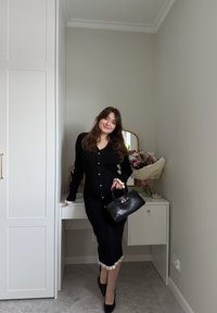 Jeune femme en robe noire et talons, tenant un sac à main noir, appuyée contre un bureau blanc avec des fleurs et un miroir dans une pièce aux tons neutres.