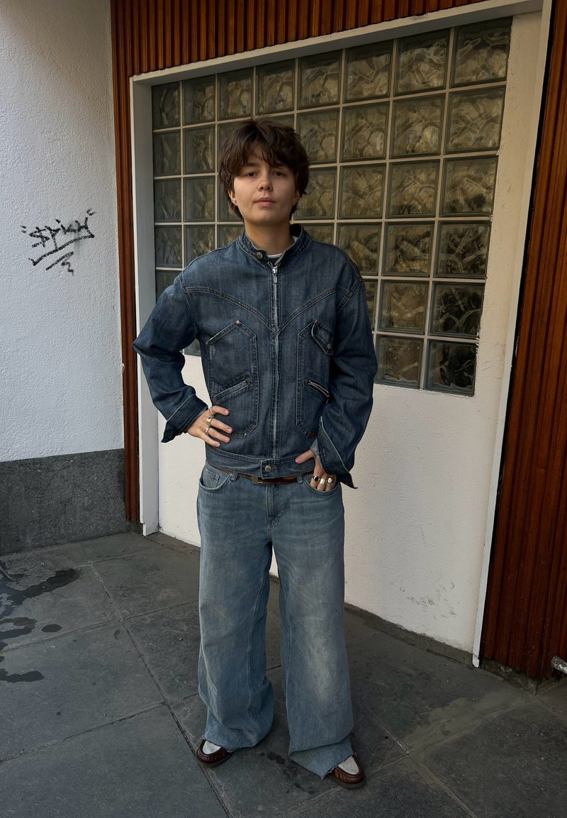 Personne aux cheveux courts portant une veste en jean et un jean à jambes larges, debout avec une main sur la hanche près d'une fenêtre en blocs de verre.