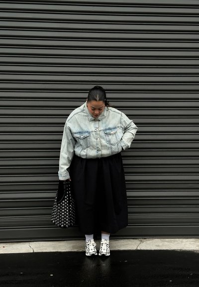 Camisa de mezclilla con botones y lavado claro, falda midi negra, bolso negro con tachuelas, calcetines blancos y zapatillas plateadas, sobre un fondo gris con rayas.